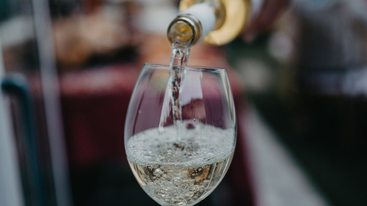 Pale white wine streaming from an unlabelled bottle neck into a clear stemmed glass, with a soft-focus garden behind.