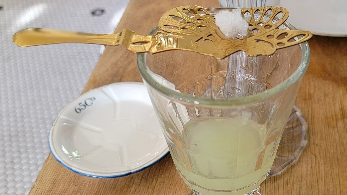 A slotted gilt absinthe spoon with a sugar cube resting across a stemmed glass of pale, cloudy green absinthe on a wooden bar.