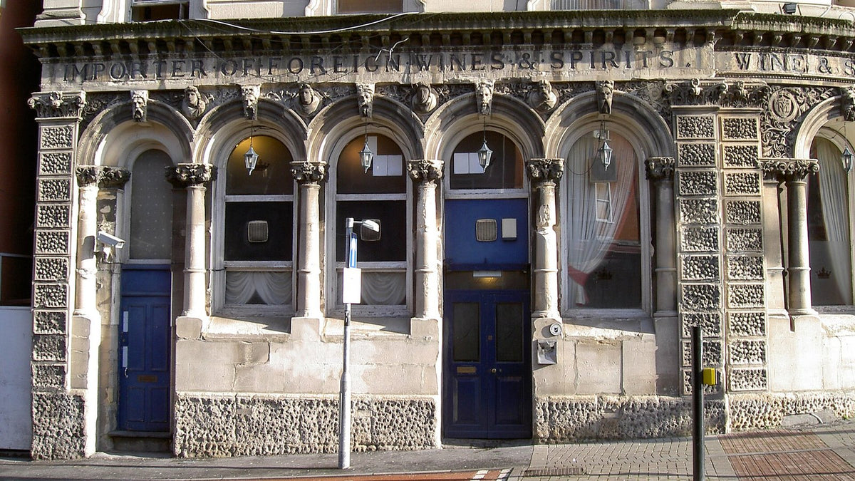 An ornate Victorian stone pub frontage carved "Importer of Foreign Wines & Spirits", with arched windows and blue doors.