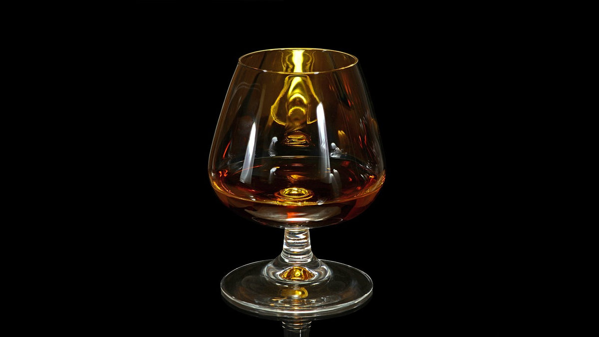 A brandy snifter holding a dark amber aged spirit against a black background.