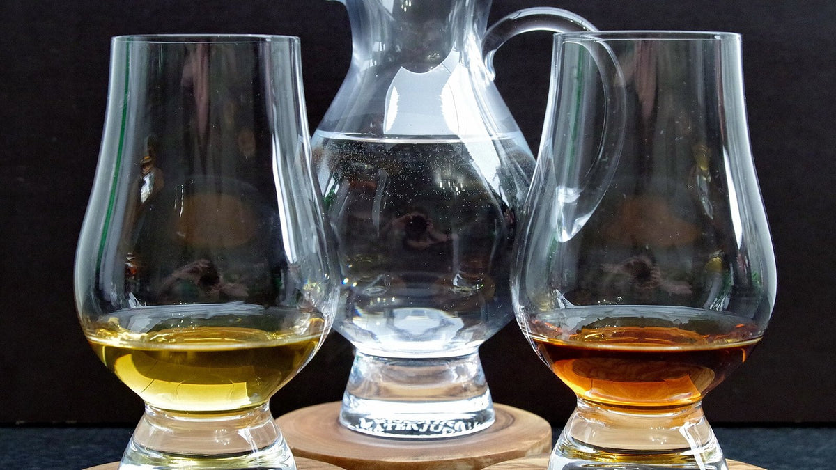 Two tulip-shaped whisky nosing glasses holding pale and amber spirit, with a small water jug between them.