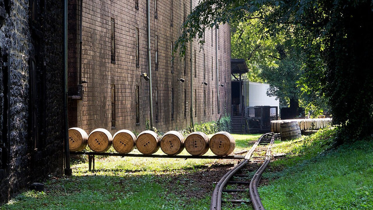 A row of barrels on a rail track beside a tall stone-and-brick bourbon warehouse in Kentucky.