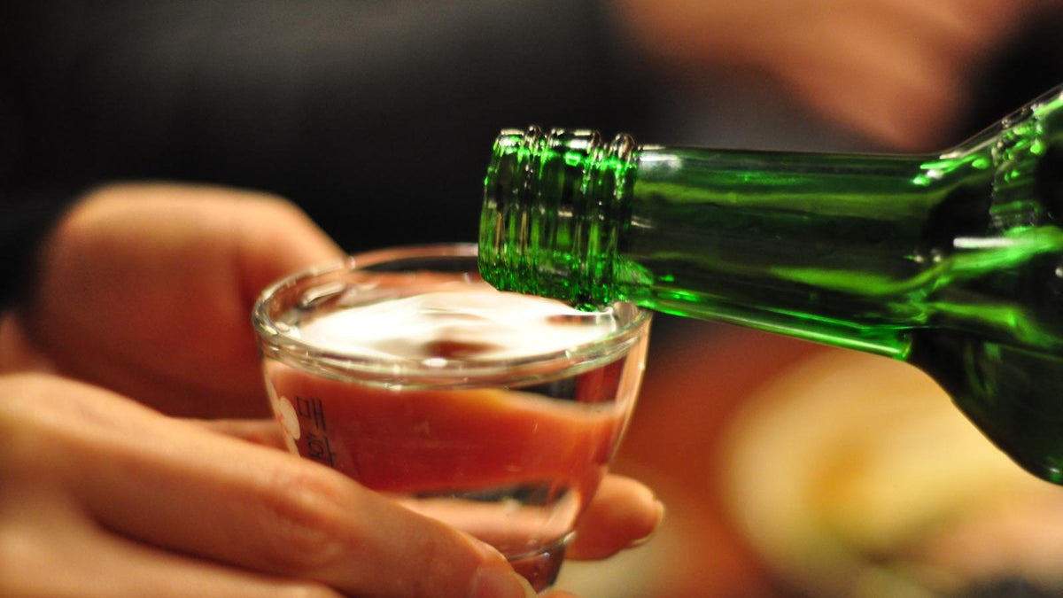 A green glass bottle with no visible label pouring soju into a small glass held in a hand.