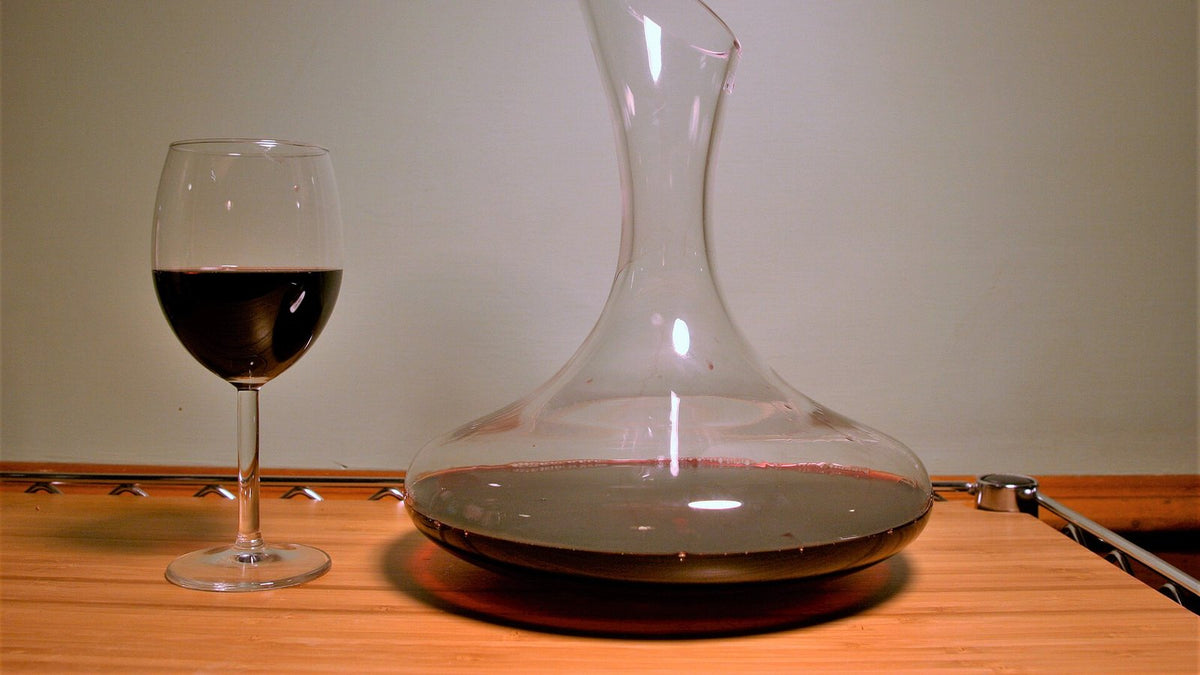 A glass of red wine beside a wide glass decanter partly filled with red wine on a wooden table.