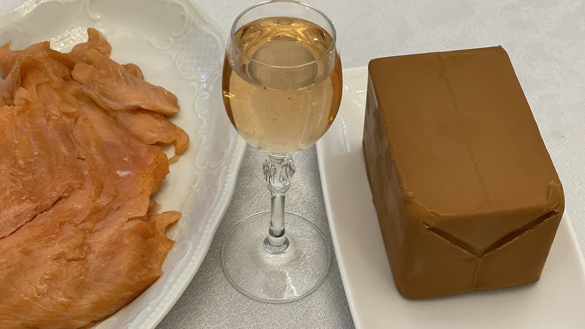 A stemmed glass of pale golden aquavit between a plate of cured salmon and a block of brown Norwegian cheese.