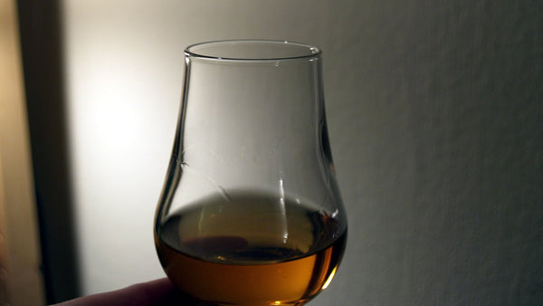A hand holding a tulip-shaped tasting glass of amber whisky against a pale wall.