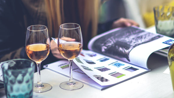 Two stemmed glasses of pale salmon-pink rosé on a white café table beside an open magazine in daylight.