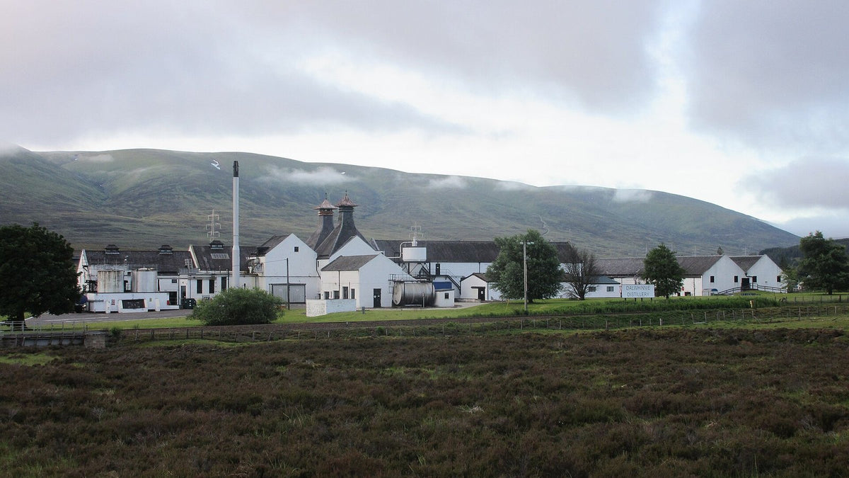 A white-walled distillery with pagoda roofs below a cloud-capped Highland hillside, with heather in front.