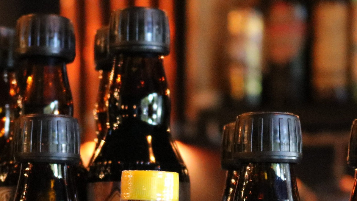 The capped tops of a crowd of small cocktail-bitters bottles on a bar counter, with warm bar lights behind.