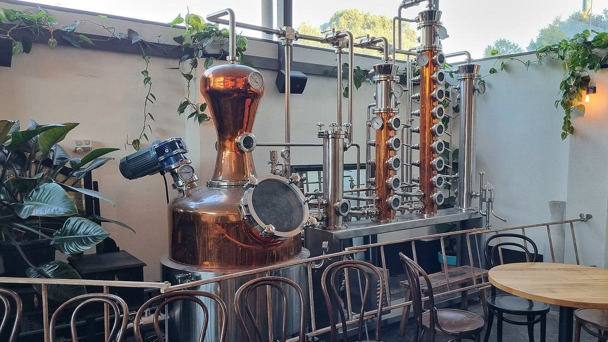 A copper pot still joined to two glass-windowed rectifying columns, installed behind a pub seating area.