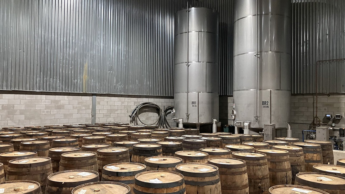 Rows of upright whiskey barrels in front of two tall stainless-steel blending tanks.
