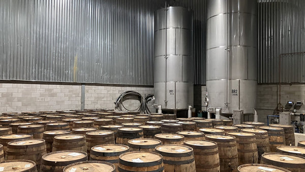 Rows of upright whiskey barrels in front of two tall stainless-steel blending tanks.