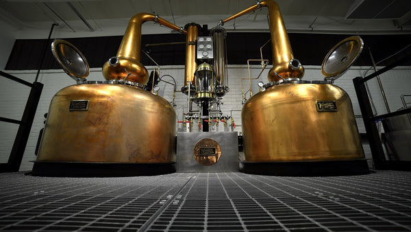Two copper pot stills seen head-on across a steel grate floor in a modern American craft distillery.