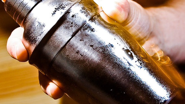 Two hands gripping a frosted steel cobbler shaker mid-shake, the far end blurred with motion.
