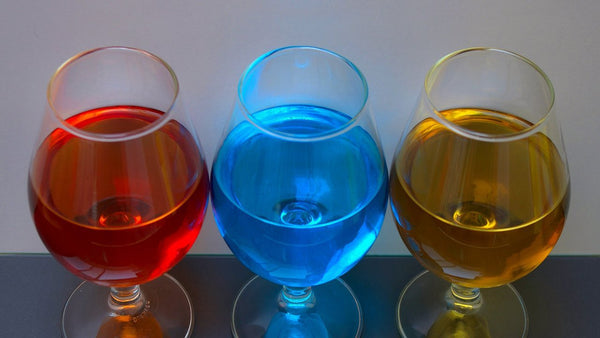 Three snifter glasses of bright red, blue and amber liqueur side by side against a pale wall.
