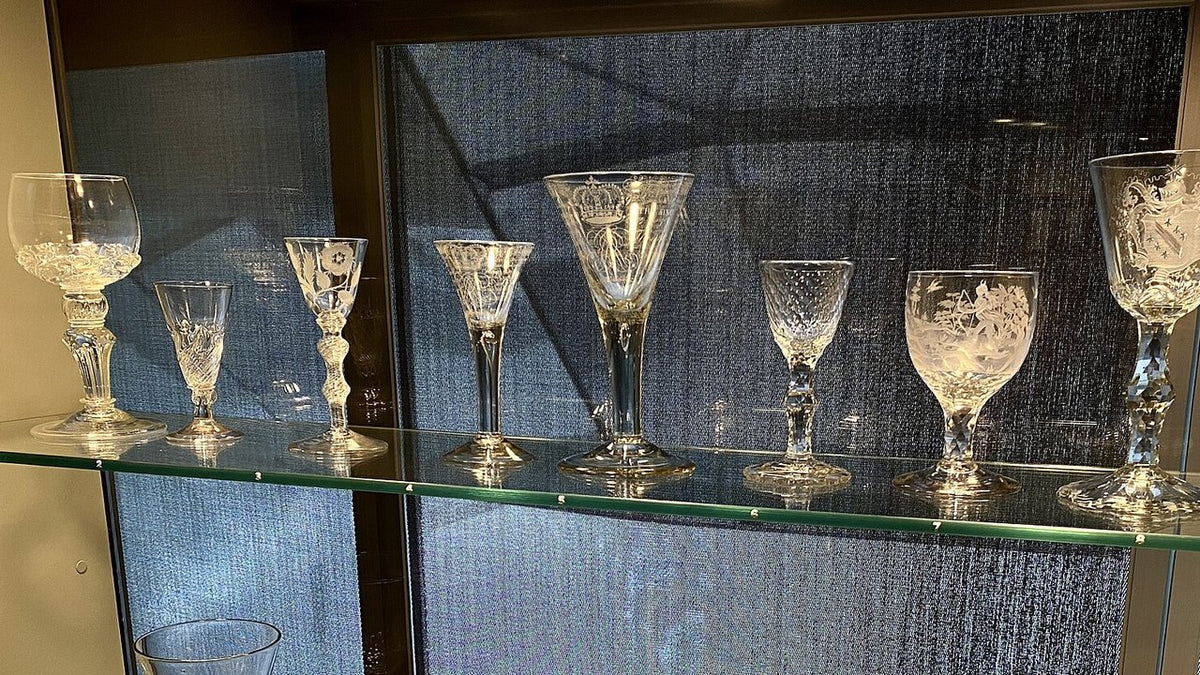 A row of empty stemmed drinking glasses in different bowl and stem shapes on a glass shelf.