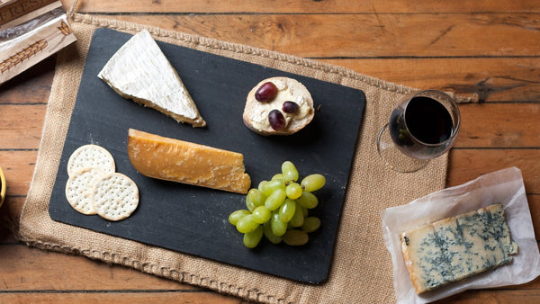 Overhead view of a slate cheese board with brie, hard cheese, grapes and crackers beside a glass of red wine and a wedge of blue cheese.