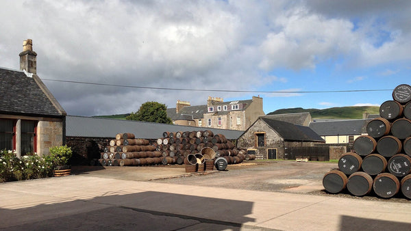 A distillery courtyard stacked with whisky casks, with Campbeltown houses and green hills behind.