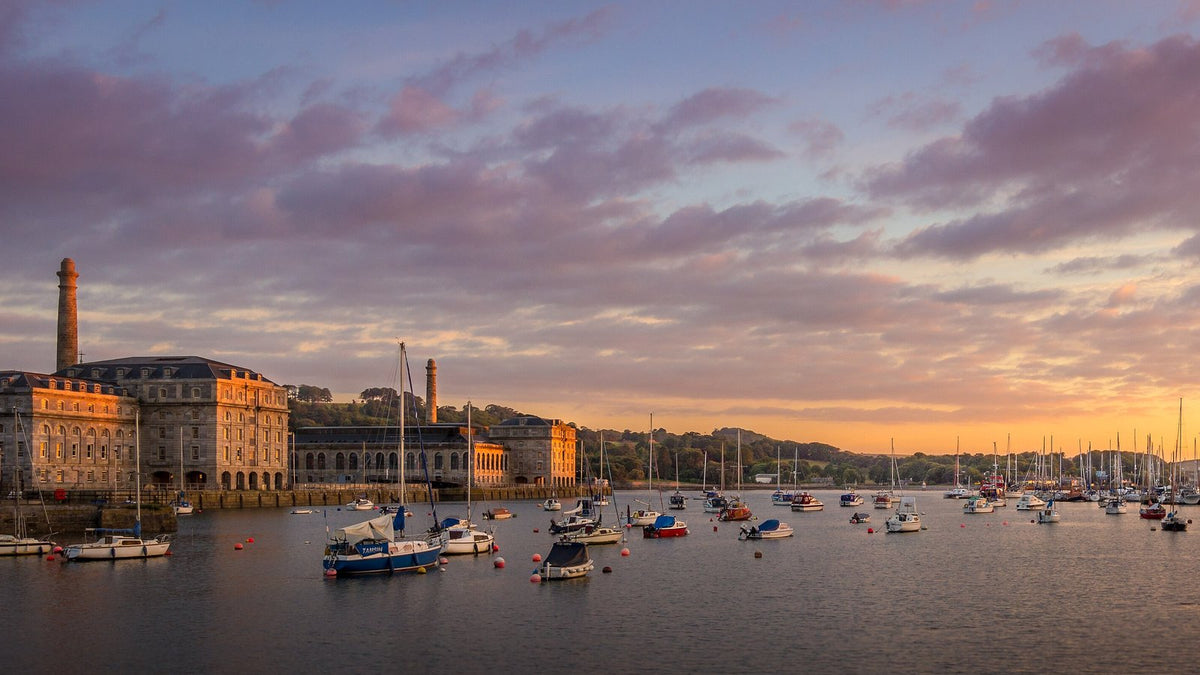 The stone buildings of the Royal William Yard, the Royal Navy's former victualling yard at Plymouth, across a harbour of moored boats at sunset.