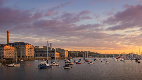 The stone buildings of the Royal William Yard, the Royal Navy's former victualling yard at Plymouth, across a harbour of moored boats at sunset.
