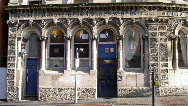 An ornate Victorian stone pub frontage carved "Importer of Foreign Wines & Spirits", with arched windows and blue doors.
