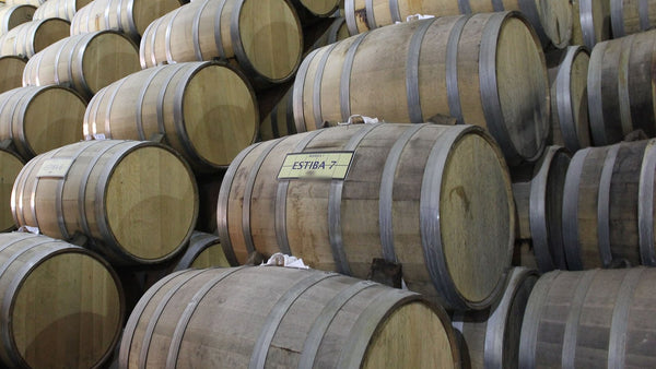 Stacked pale oak barrels ageing tequila in a bodega, one marked with a plain stack-number sign.