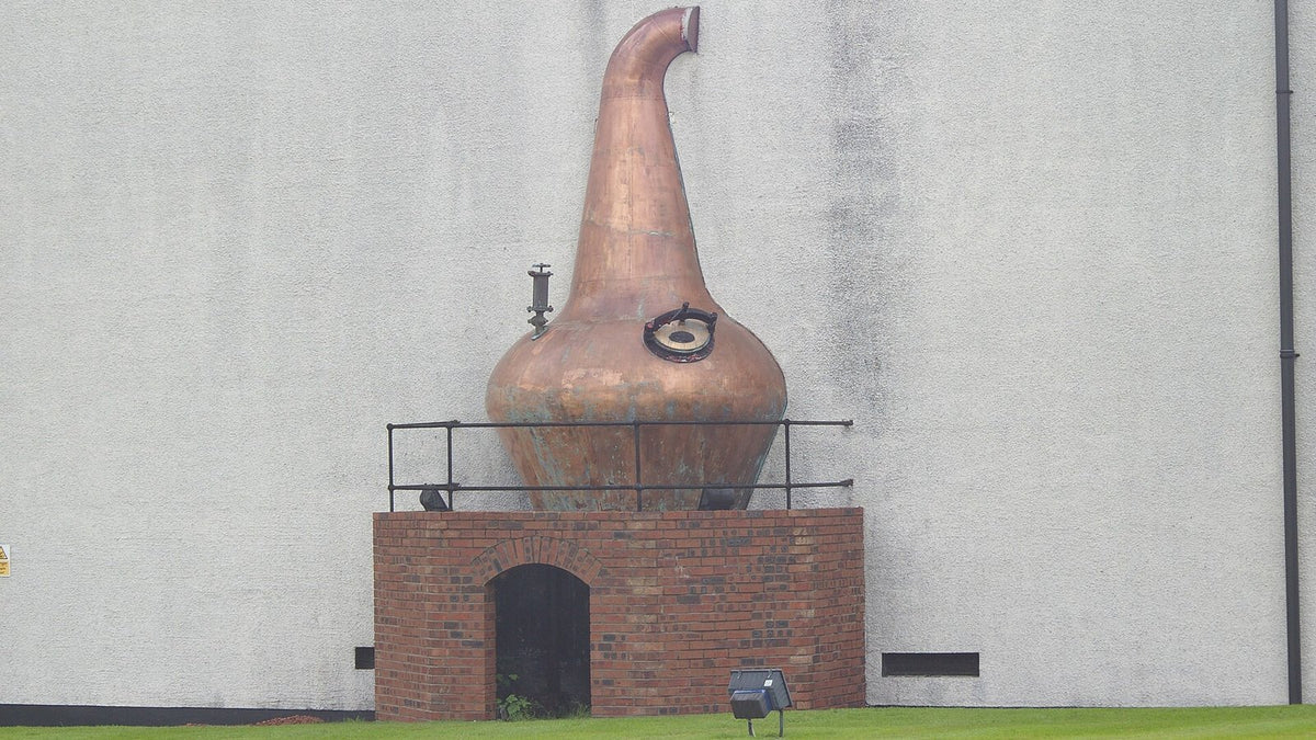 A large copper pot still displayed on a brick plinth against a white distillery wall.