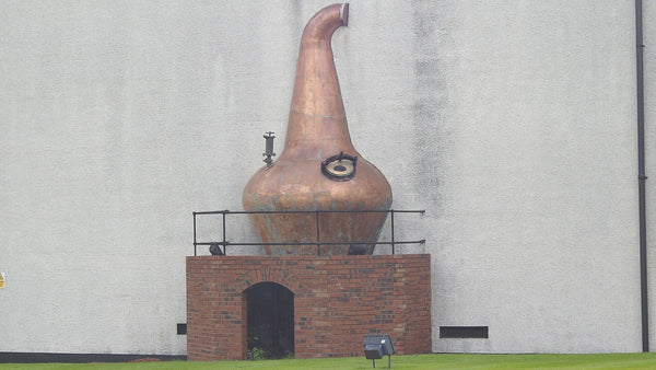 A large copper pot still displayed on a brick plinth against a white distillery wall.