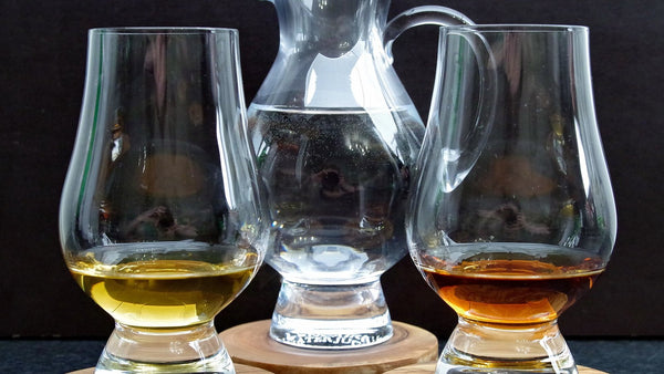 Two tulip-shaped whisky nosing glasses holding pale and amber spirit, with a small water jug between them.