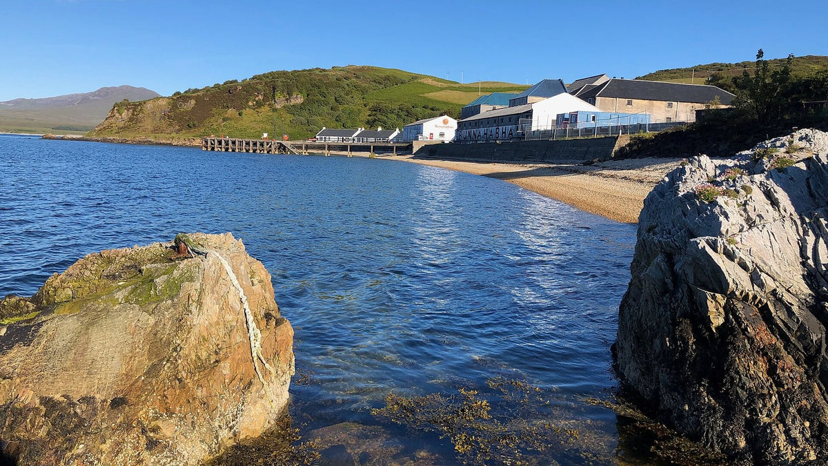 Rocks and clear seawater on an Islay shore, with a white distillery along the bay behind.