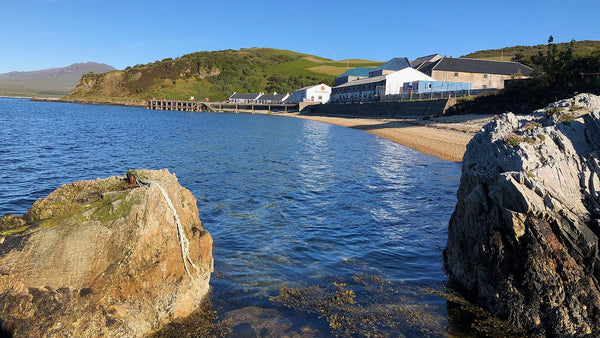 Rocks and clear seawater on an Islay shore, with a white distillery along the bay behind.