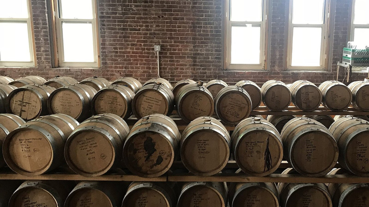 Racks of small new-oak barrels with hand-written fill notes in front of tall windows in a brick-walled craft distillery.