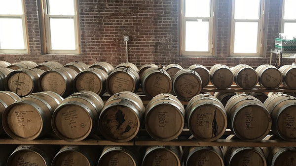 Racks of small new-oak barrels with hand-written fill notes in front of tall windows in a brick-walled craft distillery.