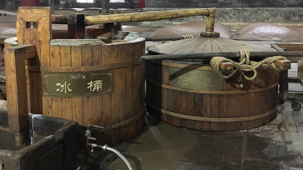 Traditional wooden baijiu still and cooling tub on a distillery floor, with mud-sealed fermentation pits behind.