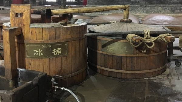 Traditional wooden baijiu still and cooling tub on a distillery floor, with mud-sealed fermentation pits behind.
