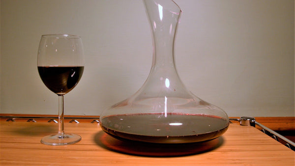 A glass of red wine beside a wide glass decanter partly filled with red wine on a wooden table.