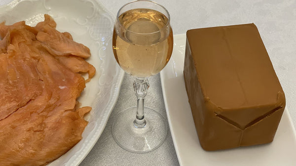 A stemmed glass of pale golden aquavit between a plate of cured salmon and a block of brown Norwegian cheese.