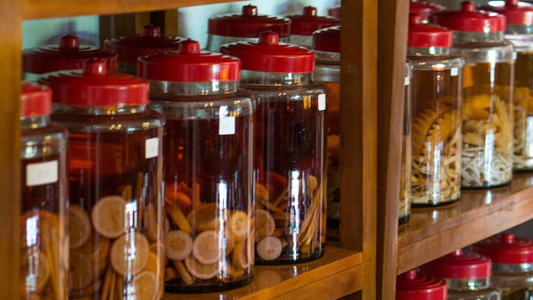 Large red-lidded glass jars of spirit infusing with dried citrus slices and spices on wooden shelves.