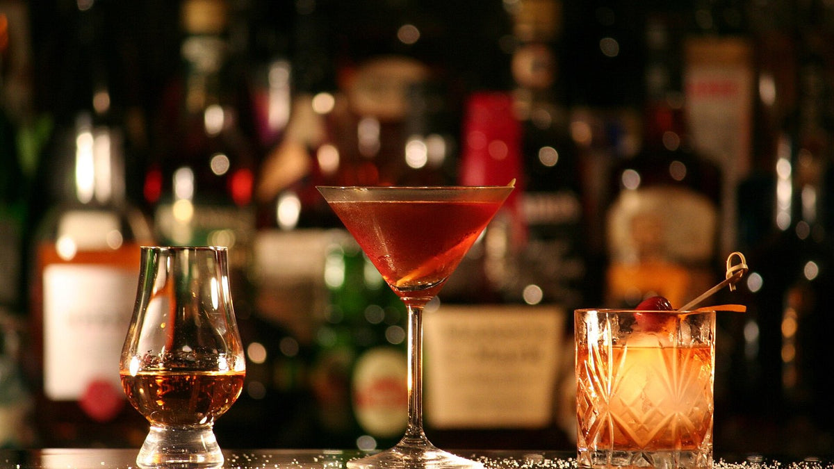 A neat rum in a nosing glass, a Manhattan in a stemmed cocktail glass and an Old Fashioned in a cut-crystal rocks glass on a bar, with bottles out of focus behind.