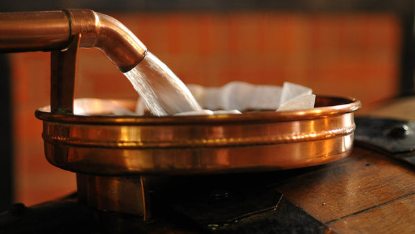Clear, freshly distilled spirit pouring from a copper still spout into a copper basin.