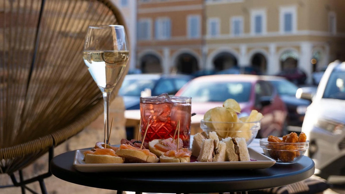 A red spritz-style drink, a glass of sparkling wine and a tray of snacks on a small outdoor café table in an Italian piazza.
