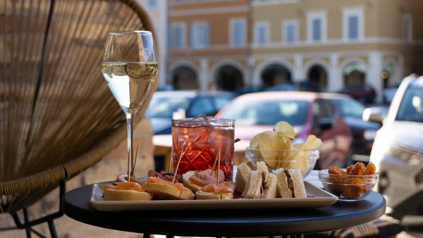 A red spritz-style drink, a glass of sparkling wine and a tray of snacks on a small outdoor café table in an Italian piazza.
